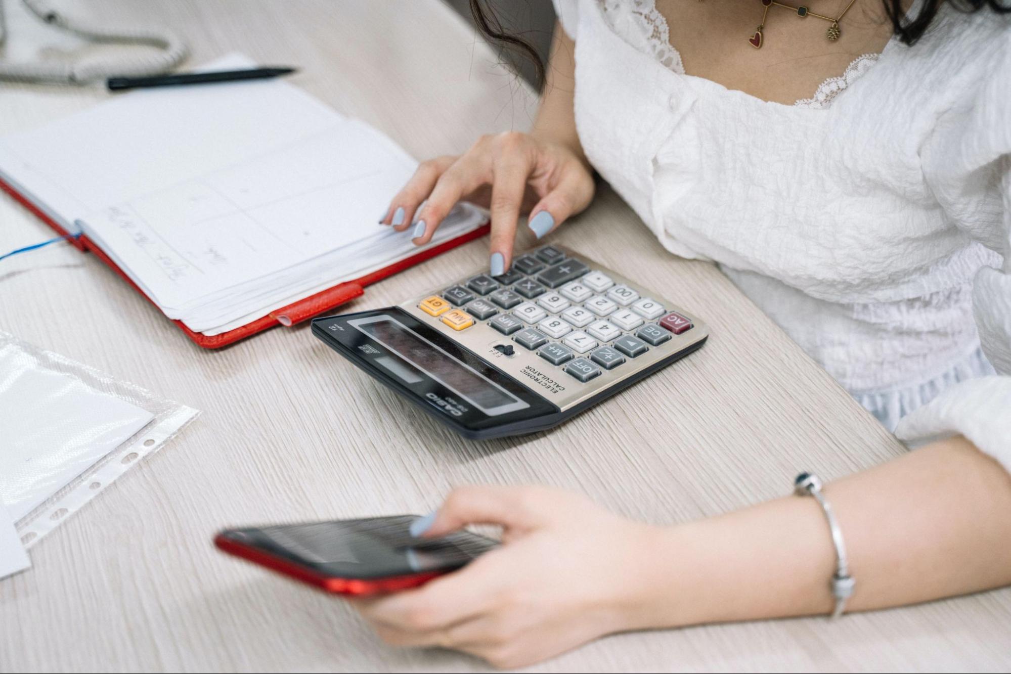 Woman sitting in front of her calculator inputting numbers. A note pad, pen and phone are around her